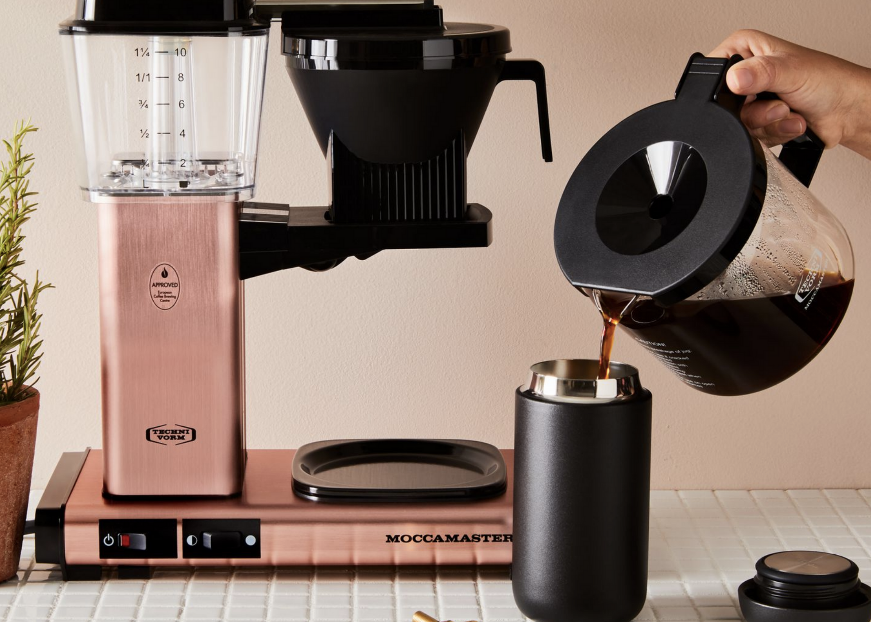 Coffee maker with a grinder and glass carafe pouring coffee into a black travel mug.