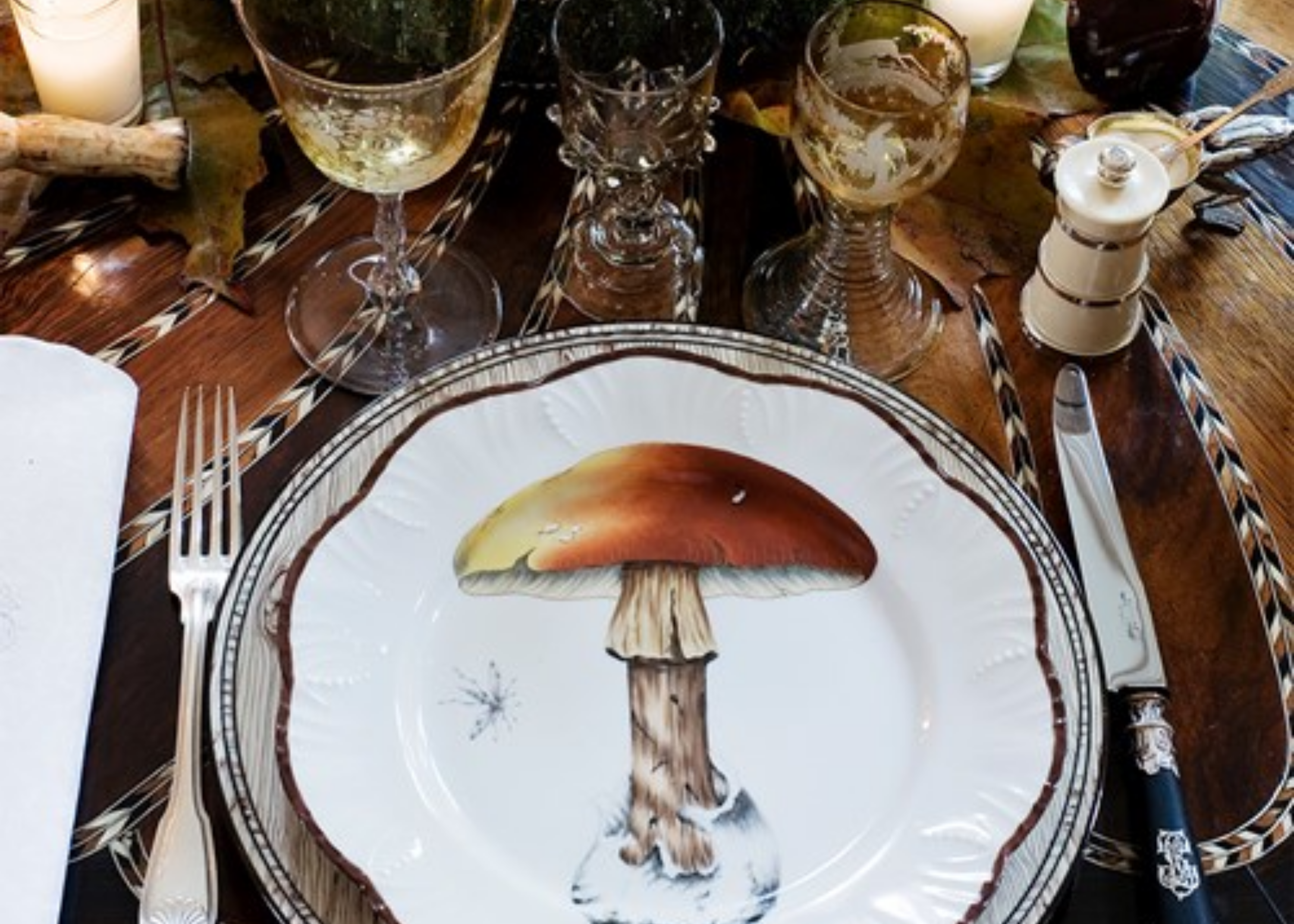 Place setting with a mushroom illustration on a plate, cutlery, patterned glasses, and a pepper mill.