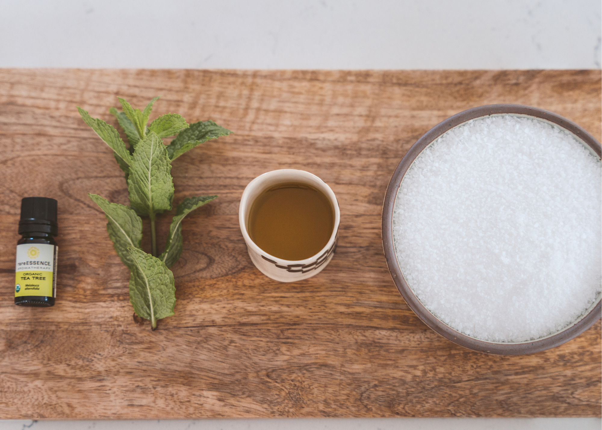 Tea tree essential oil, fresh mint leaves, a cup of liquid, and white crystals arranged on a wooden surface.