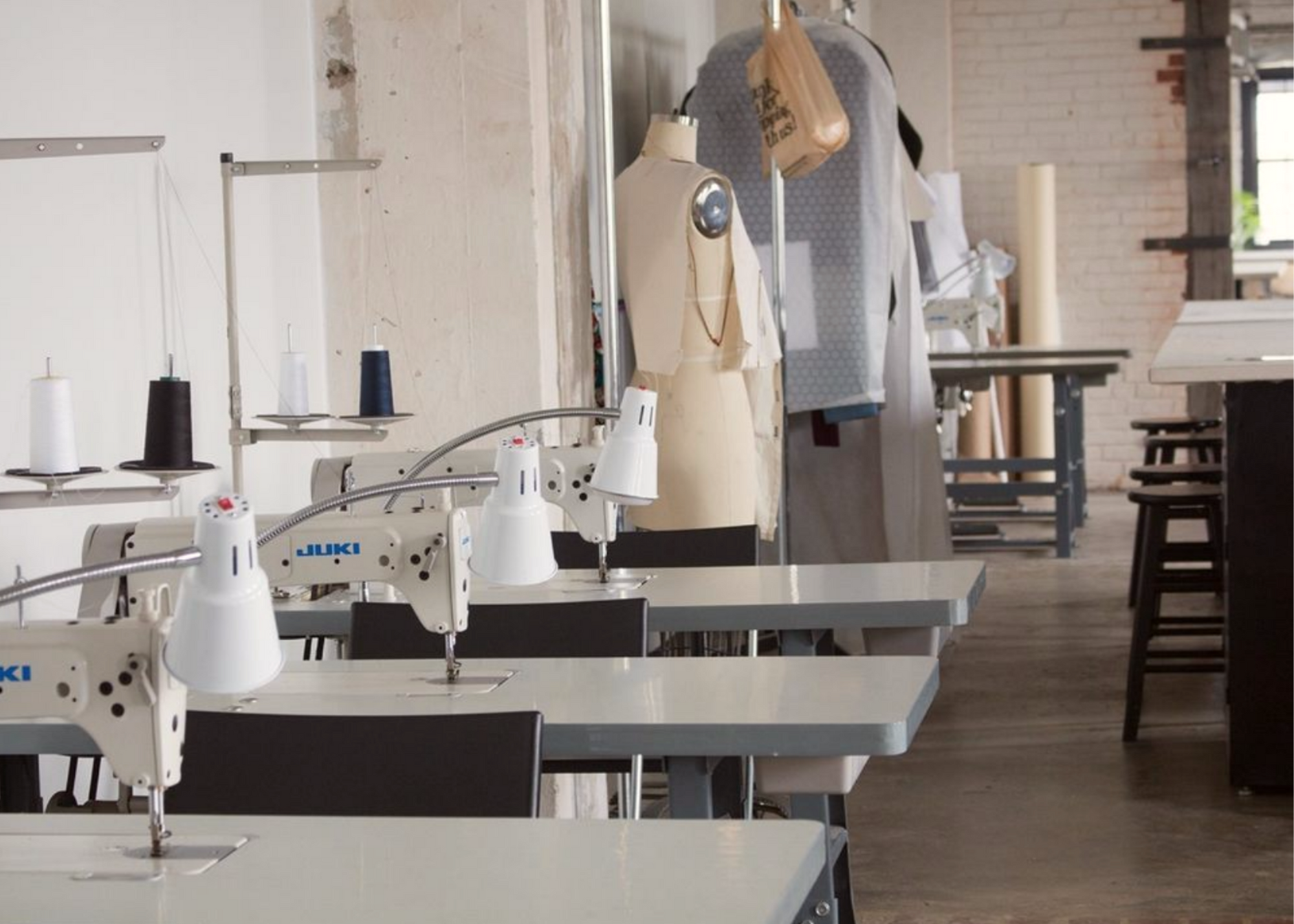 Sewing studio with industrial sewing machines, dress forms, worktables, and thread spools.