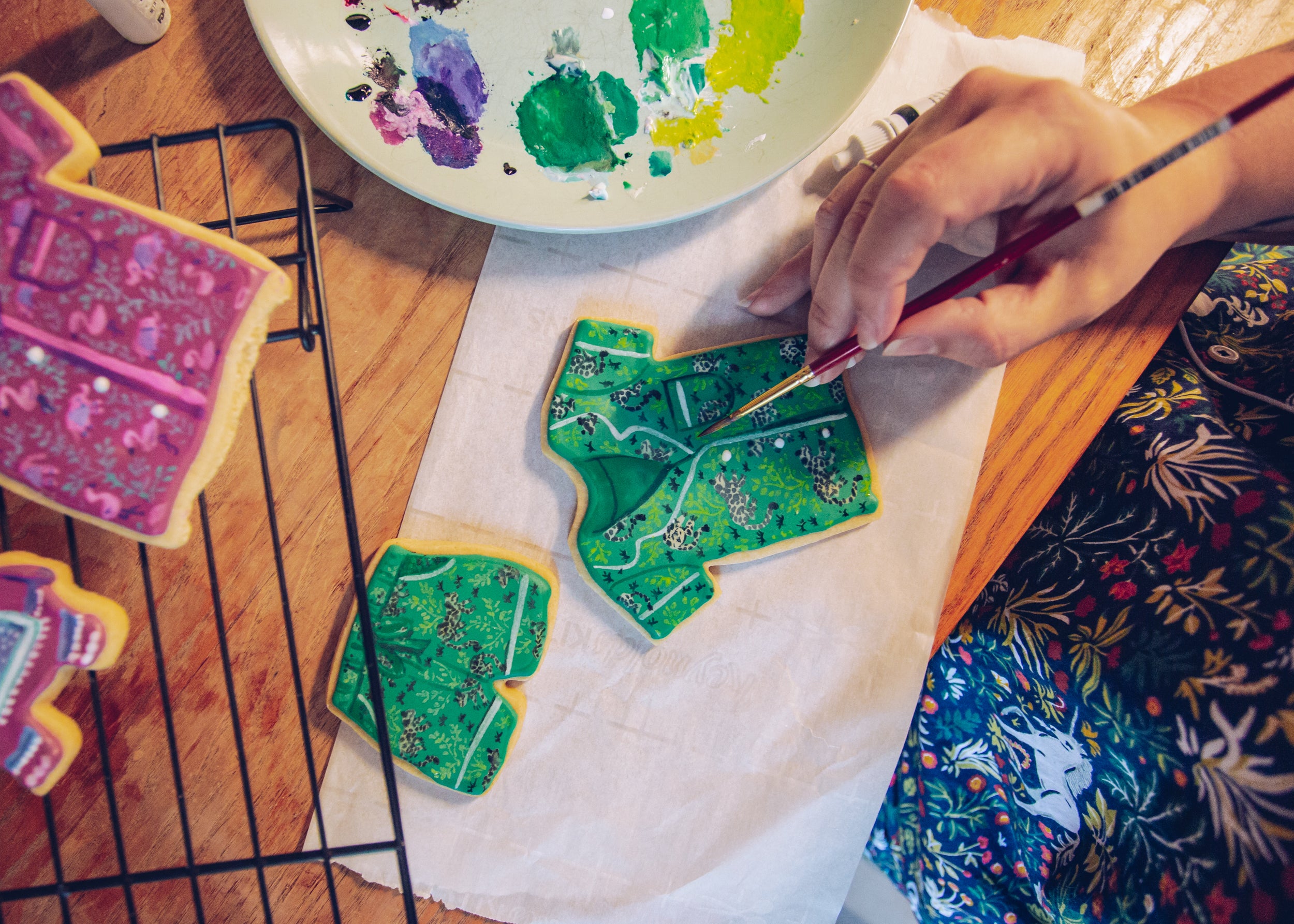 Person painting green patterned clothing-shaped cookies with a fine brush beside a paint palette.