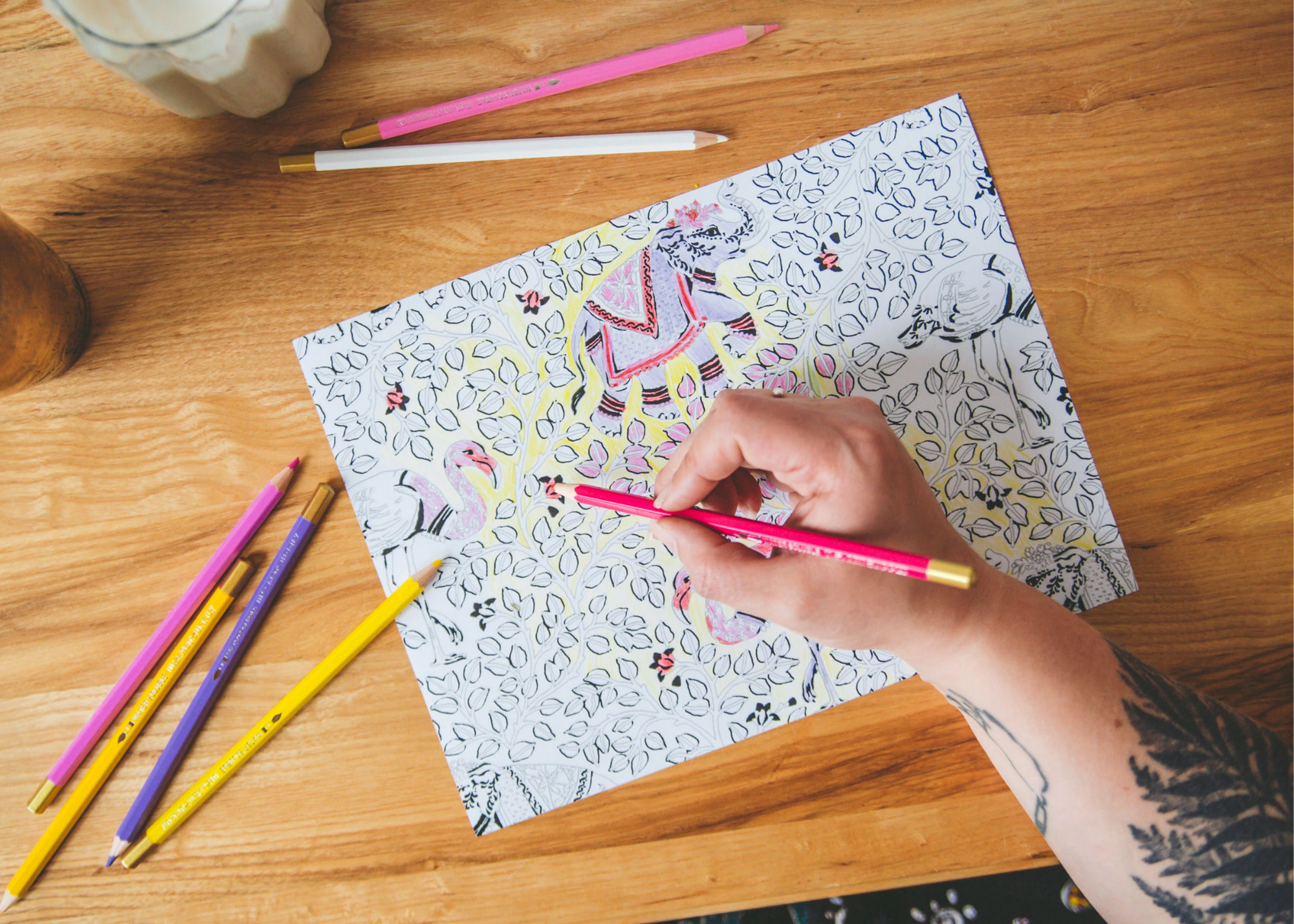 Person hand-coloring an animal-and-leaf illustration with colored pencils on a wooden table.