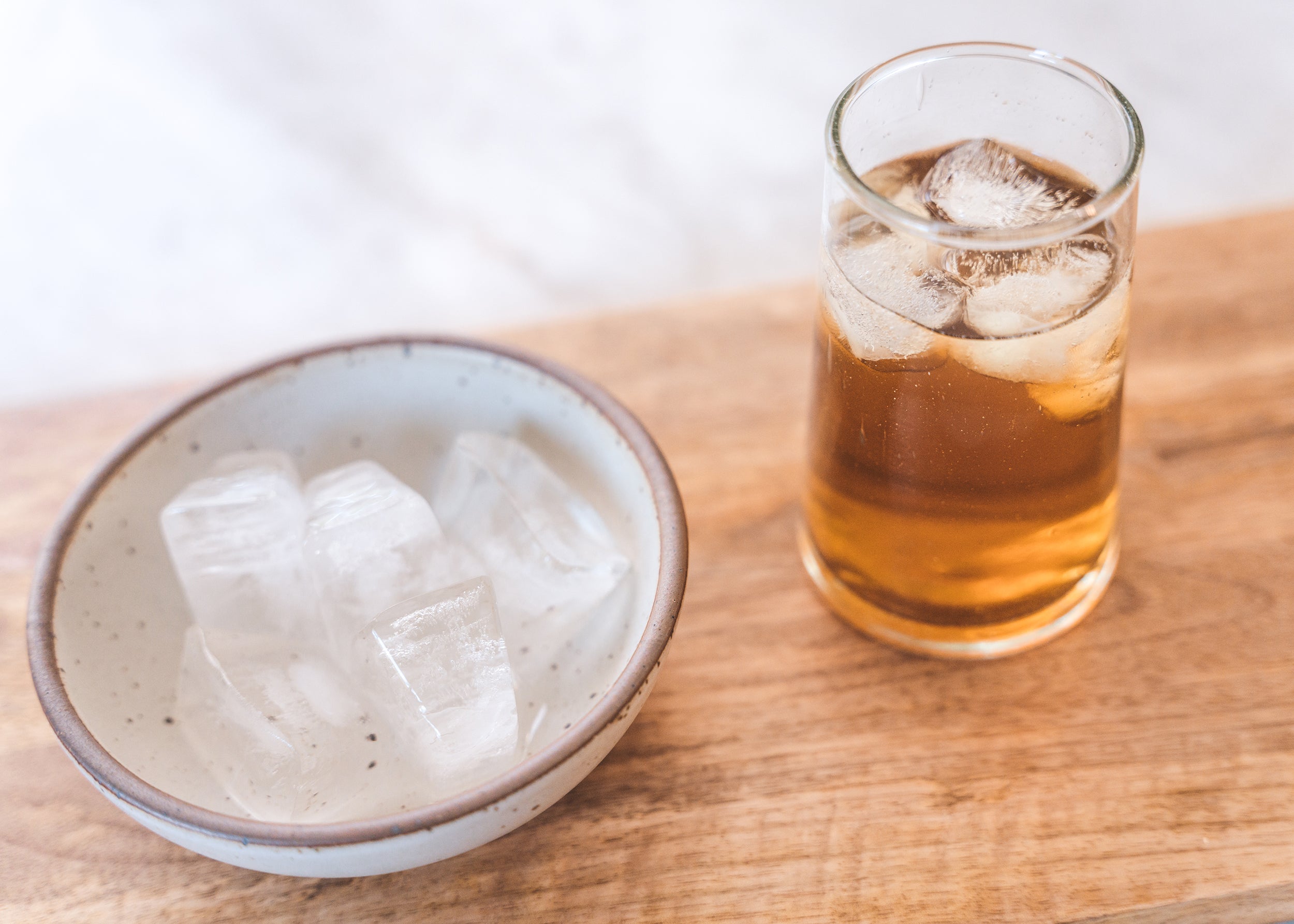 Glass of amber beverage with ice beside a bowl of ice cubes on a wooden table.