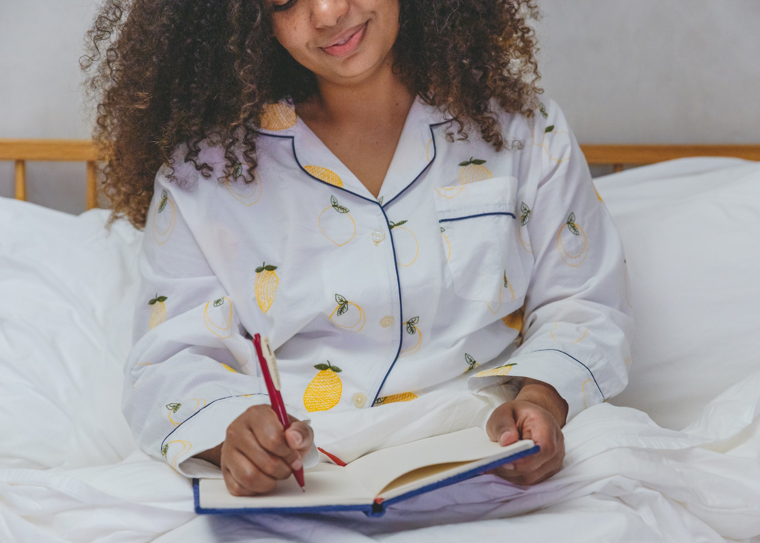Person seated on a bed writing in a notebook while wearing a light pajama top with a fruit and insect pattern.