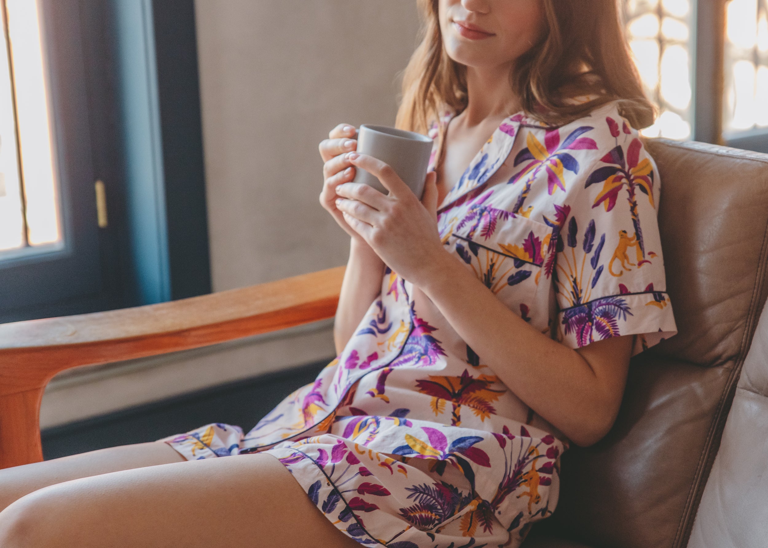 Person seated in a chair holding a mug and wearing a light patterned collared dress.