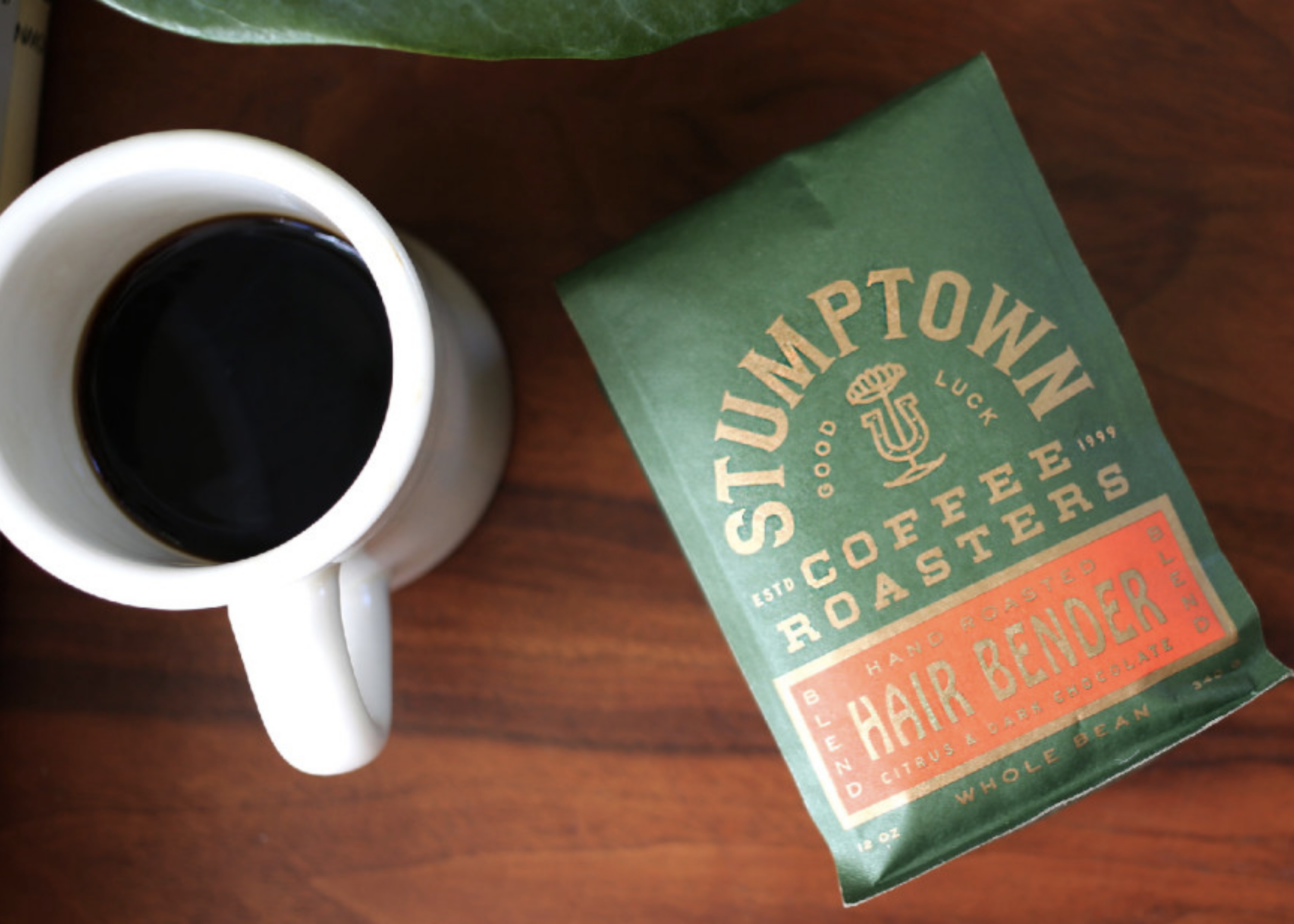 White mug of black coffee beside a green bag of Stumptown Coffee Roasters Hair Bender coffee on a wooden table.