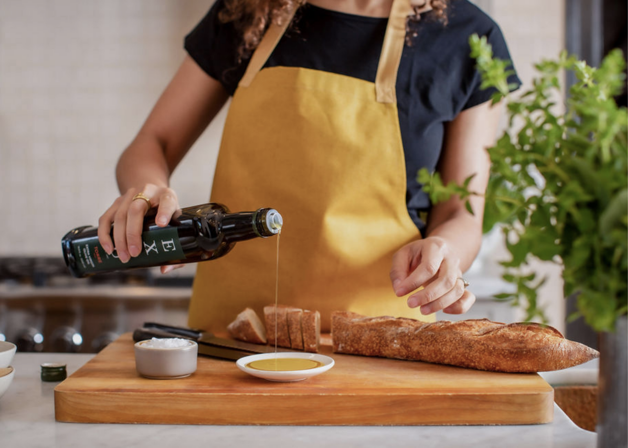 Person in a yellow apron pouring oil from a dark bottle into a dish beside sliced bread on a cutting board.