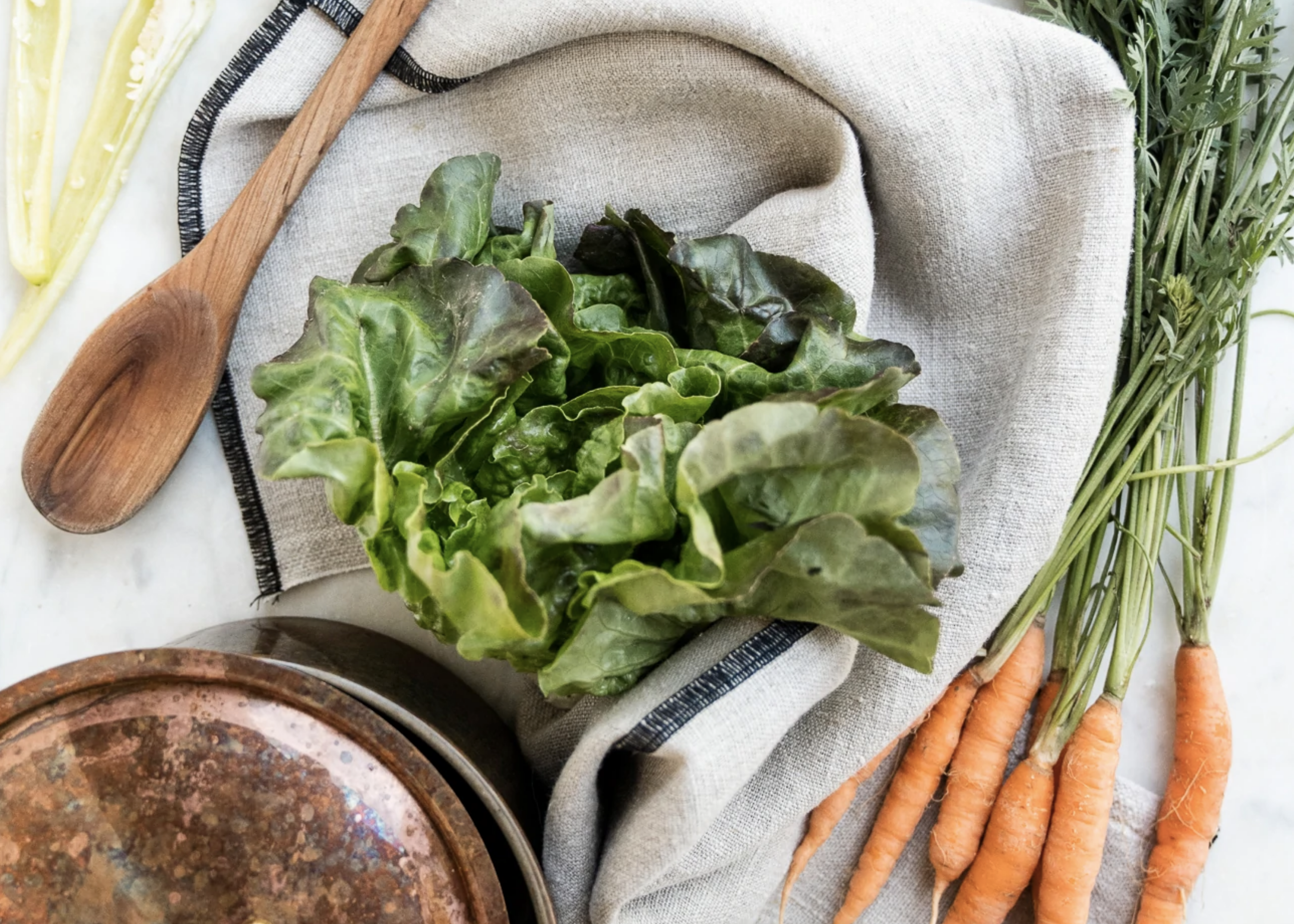 Head of green lettuce, carrots, wooden spoon, and plate arranged on a cloth-covered surface.