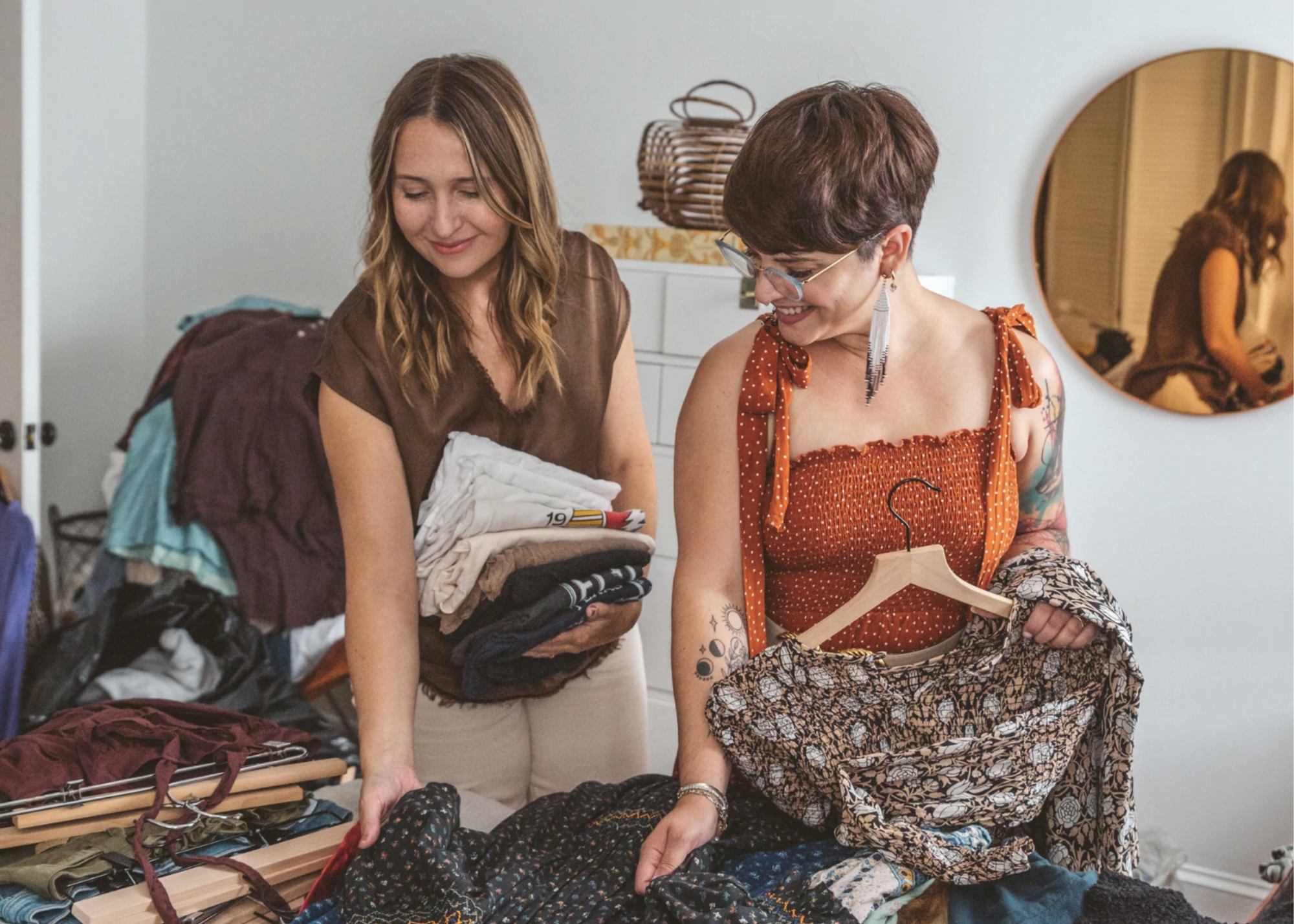 Two people sorting and holding clothing in a room with a clothing rack and mirror.