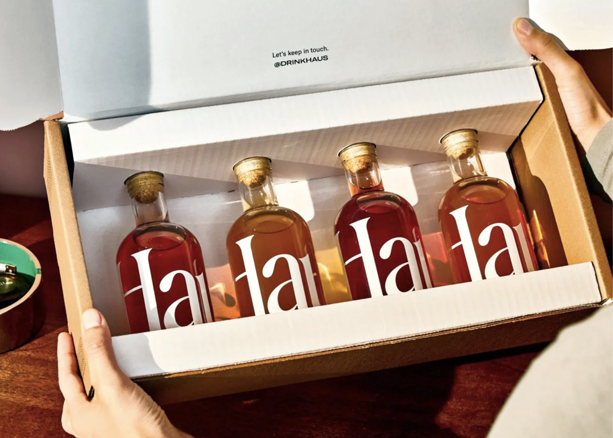 Open white box containing four glass bottles with red liquid, held by two people.