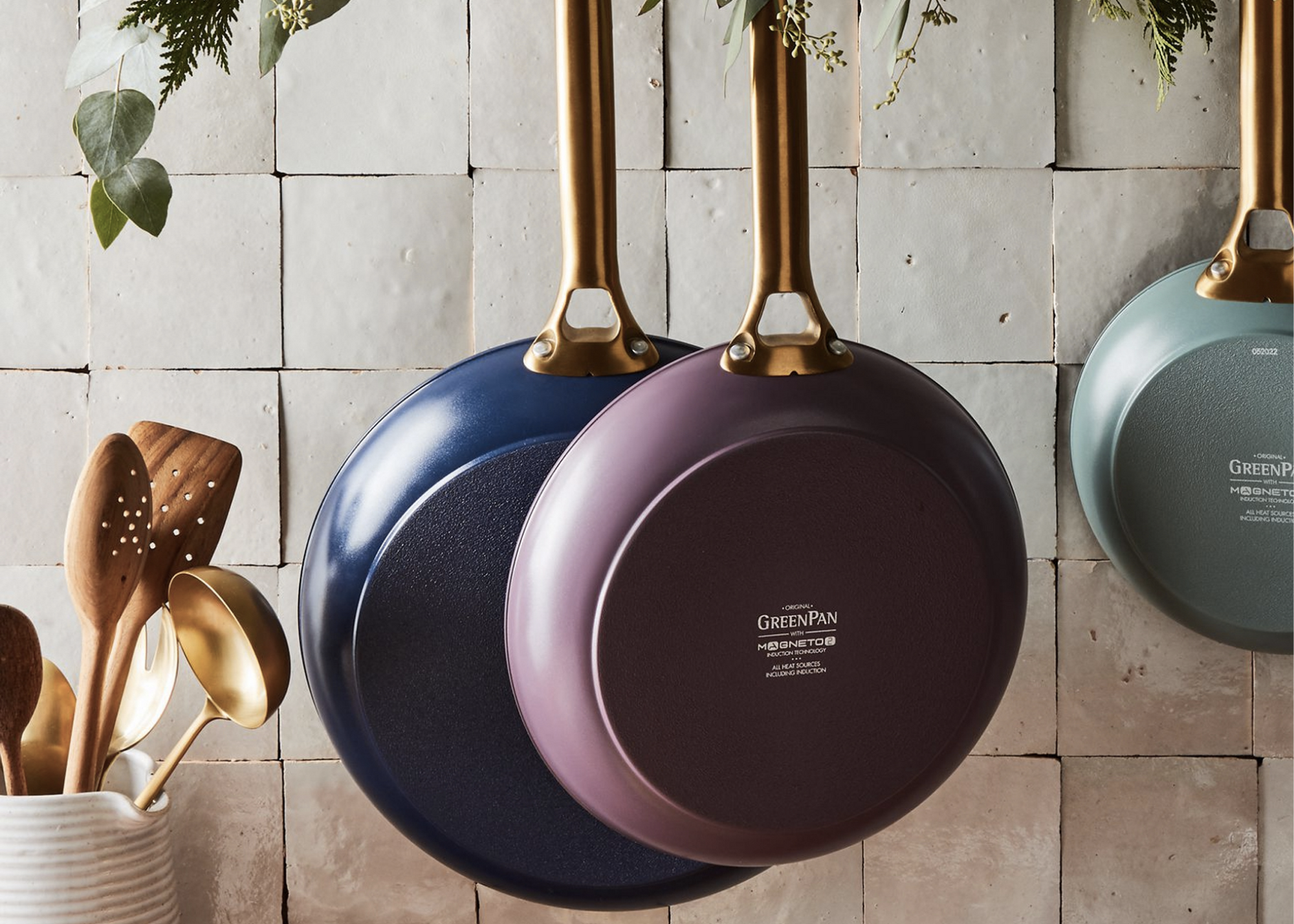 Three frying pans hanging against a tiled wall, with wooden utensils in a container at left.