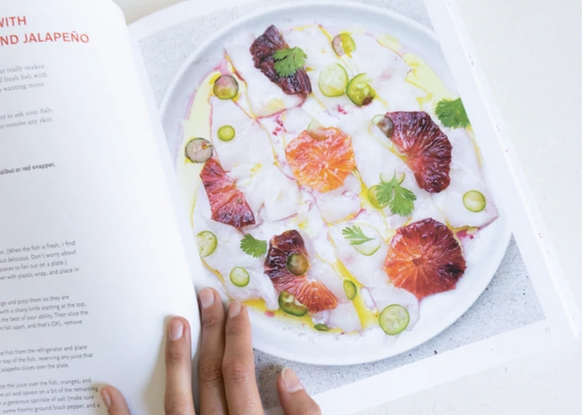 Open cookbook showing a plated dish with citrus slices, green vegetables, herbs, and a dark grilled item.