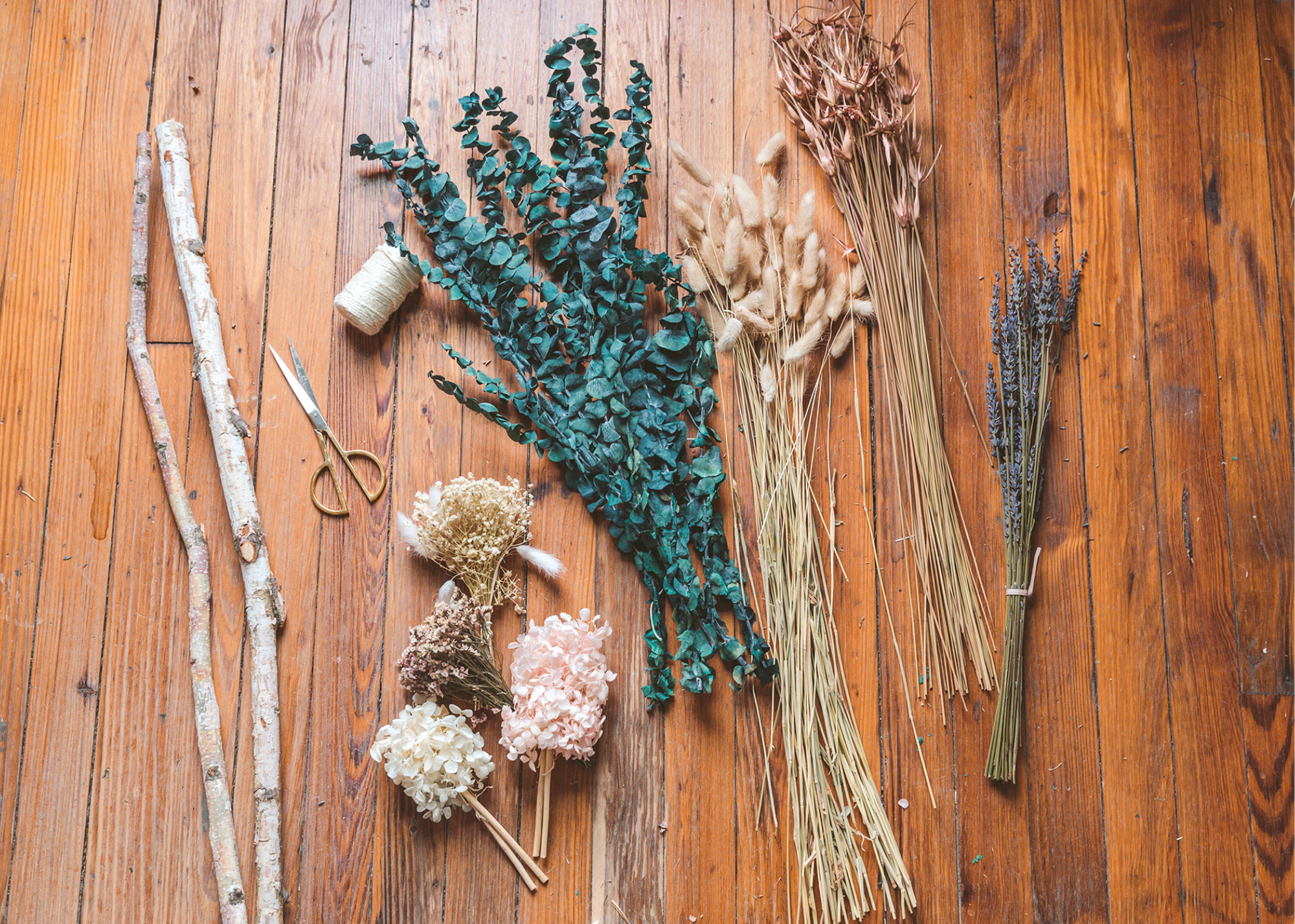 Dried flowers, plants, branches, twine, and scissors arranged on a wooden table.