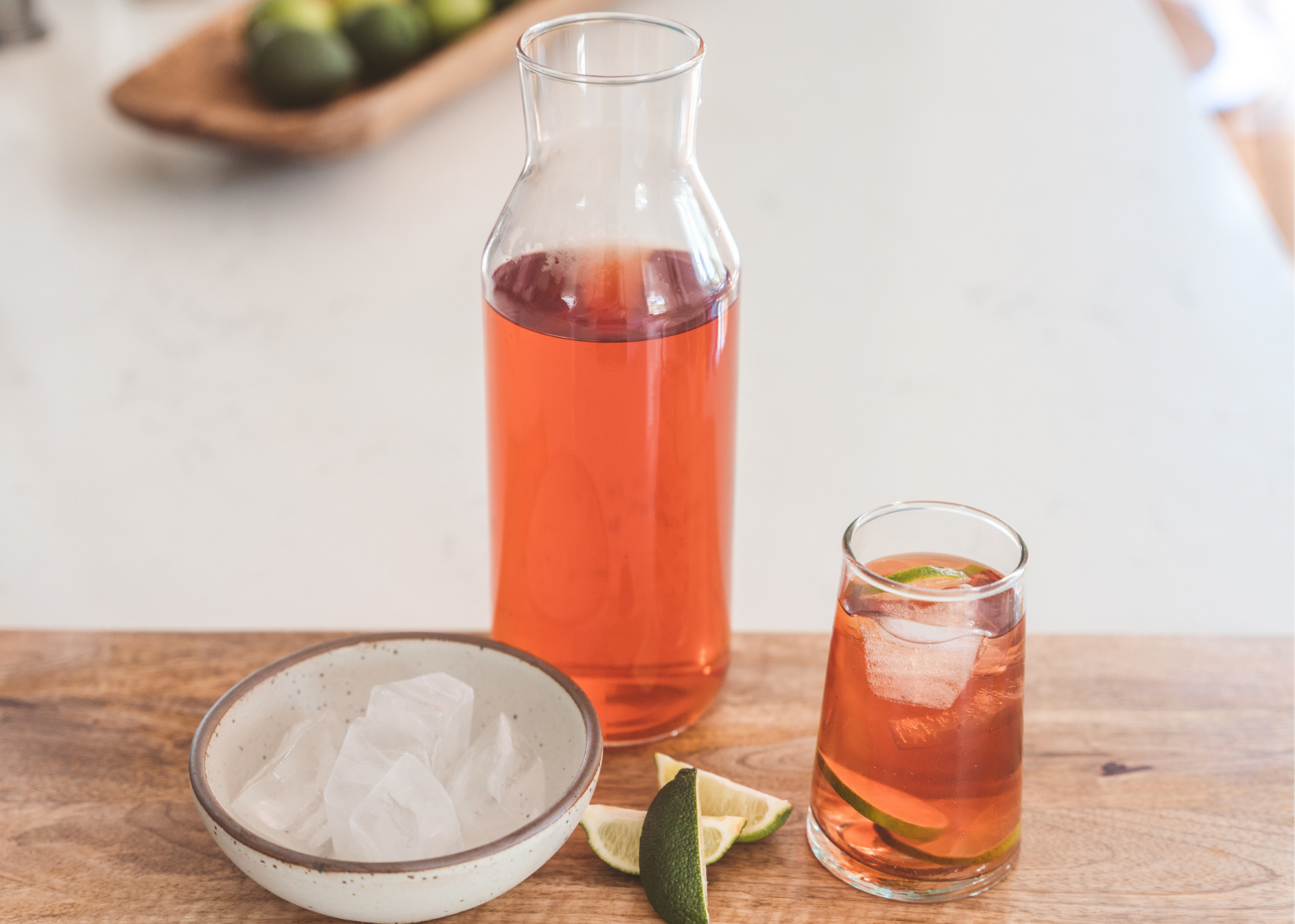 Pitcher and glass of pink beverage with ice and lime wedges beside a bowl of ice.