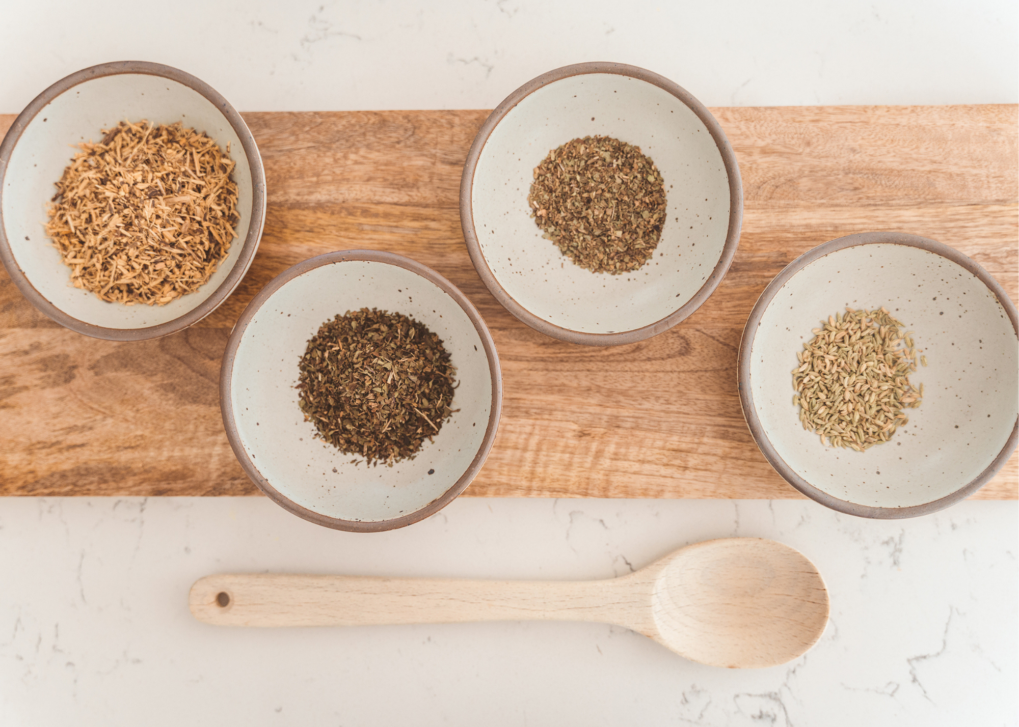 Four bowls of dried spices arranged around a wooden spoon on a wooden board.