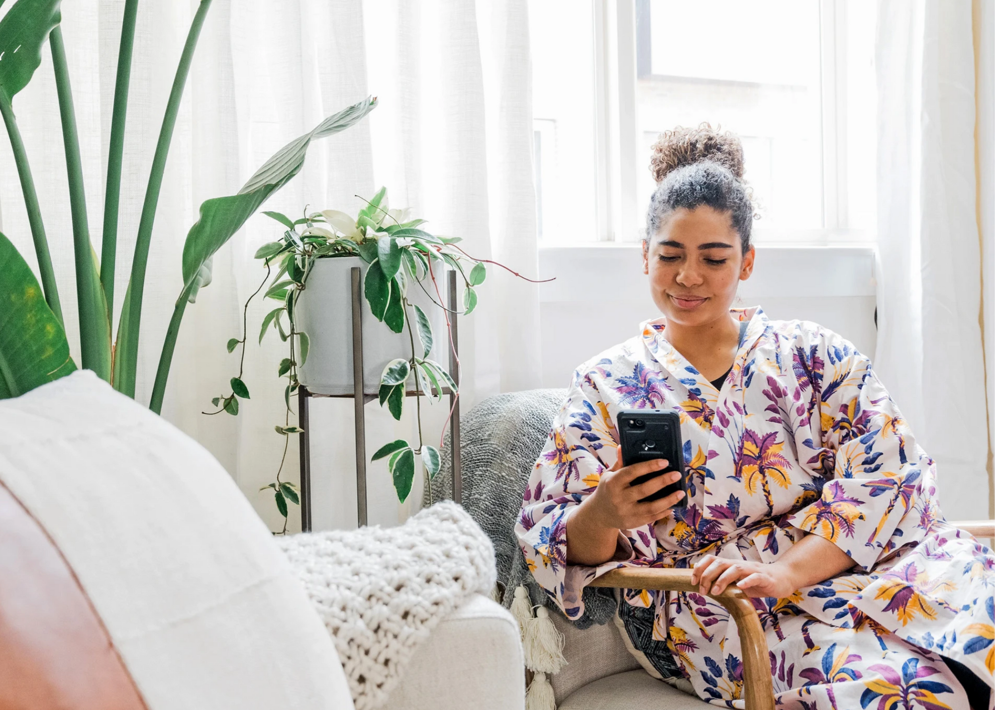 Person seated in a bright room using a smartphone beside houseplants and a chair.
