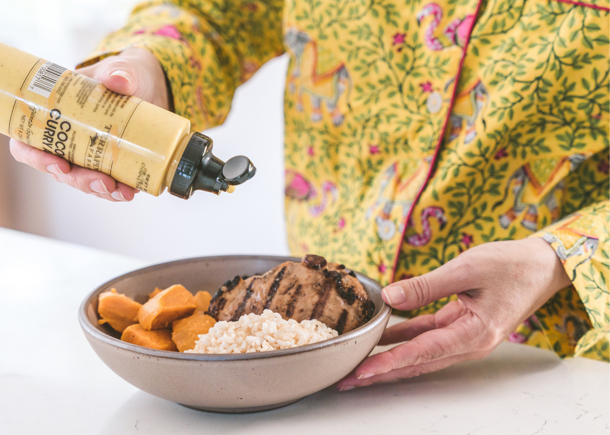 Person pours sauce from a bottle over a bowl of grilled meat, rice, and orange vegetables.