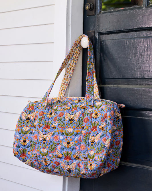Bee Tapestry quilted duffle bag hanging beside a black door, showing its blue bee-and-floral print and handles.