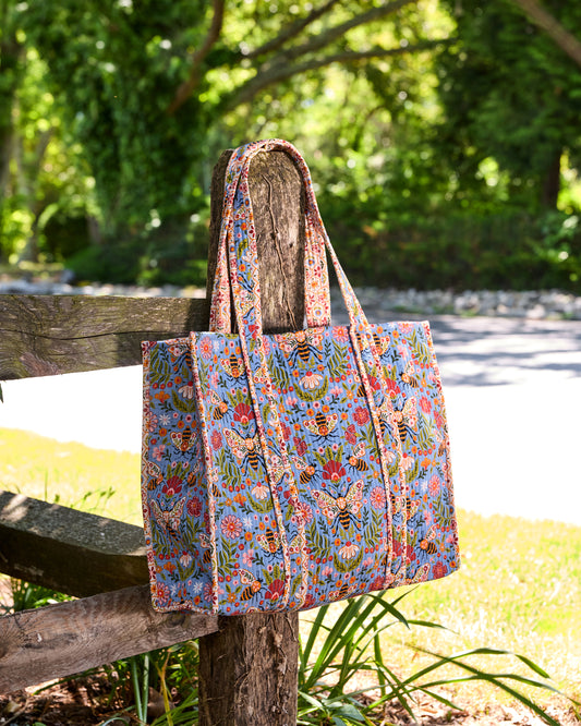 Bee Tapestry traveler tote hanging from a rustic wooden fence, shown at an angle with its blue bee-and-floral print.