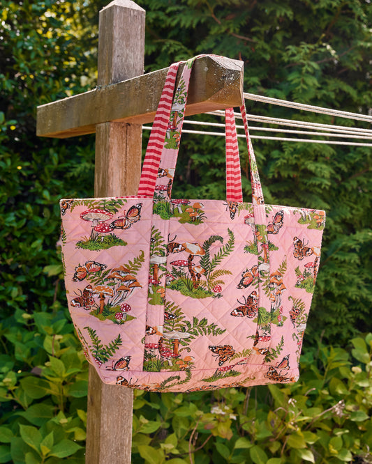 Rosewood quilted weekend bag with matching pouch hanging outdoors from a wooden structure, showing its butterfly, mushroom, and fern print.