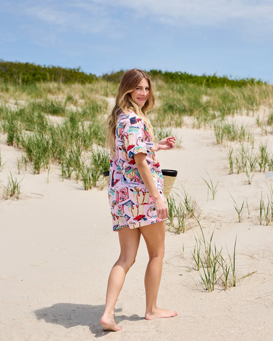 Side and back view of a person wearing a pink short-sleeve pajama set with a travel-themed print on a sandy beach.