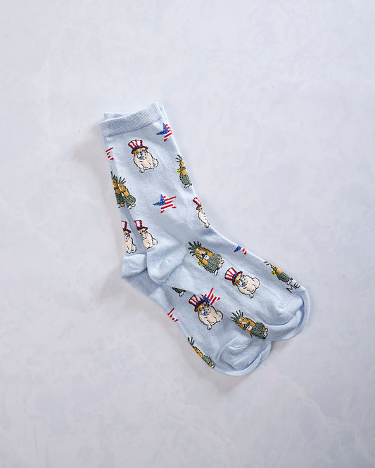 Pair of light blue crew socks with repeated patriotic dogs, pineapple-like figures, and American flag motifs.