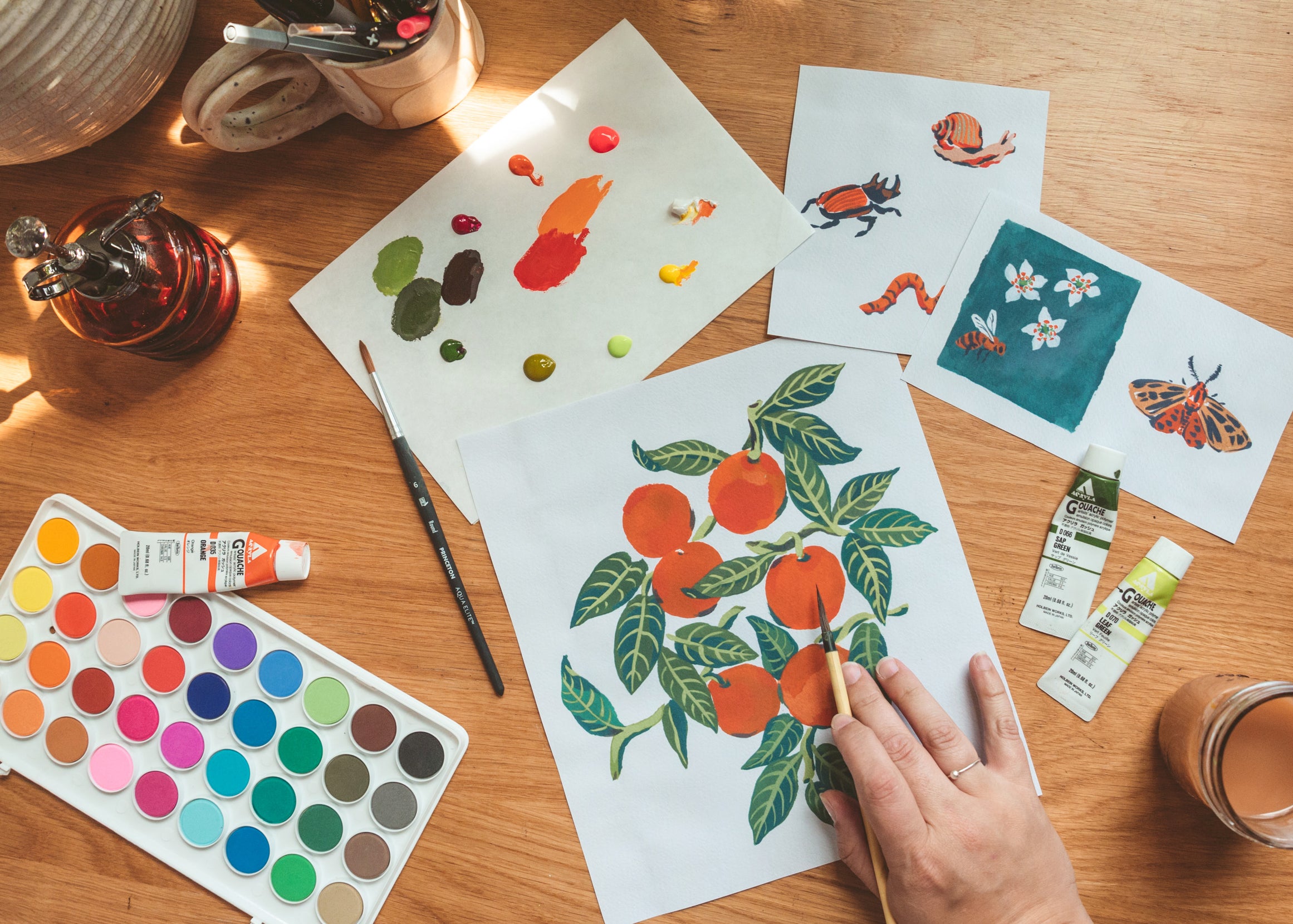 Painting workspace with orange illustrations, insect studies, paint palettes, brushes, and paint tubes.