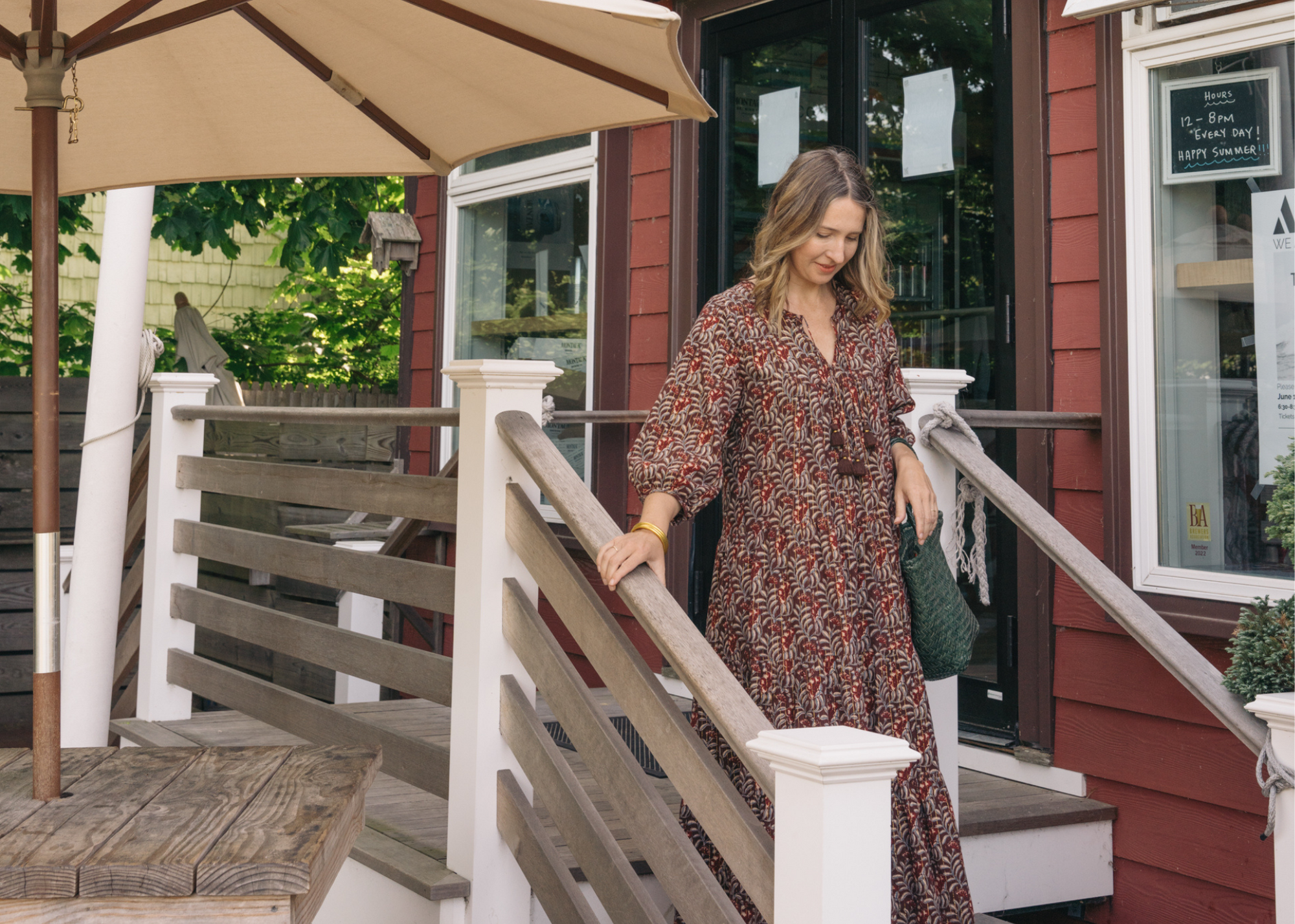 Woman in a patterned dress standing on the steps of a storefront beside an outdoor umbrella.