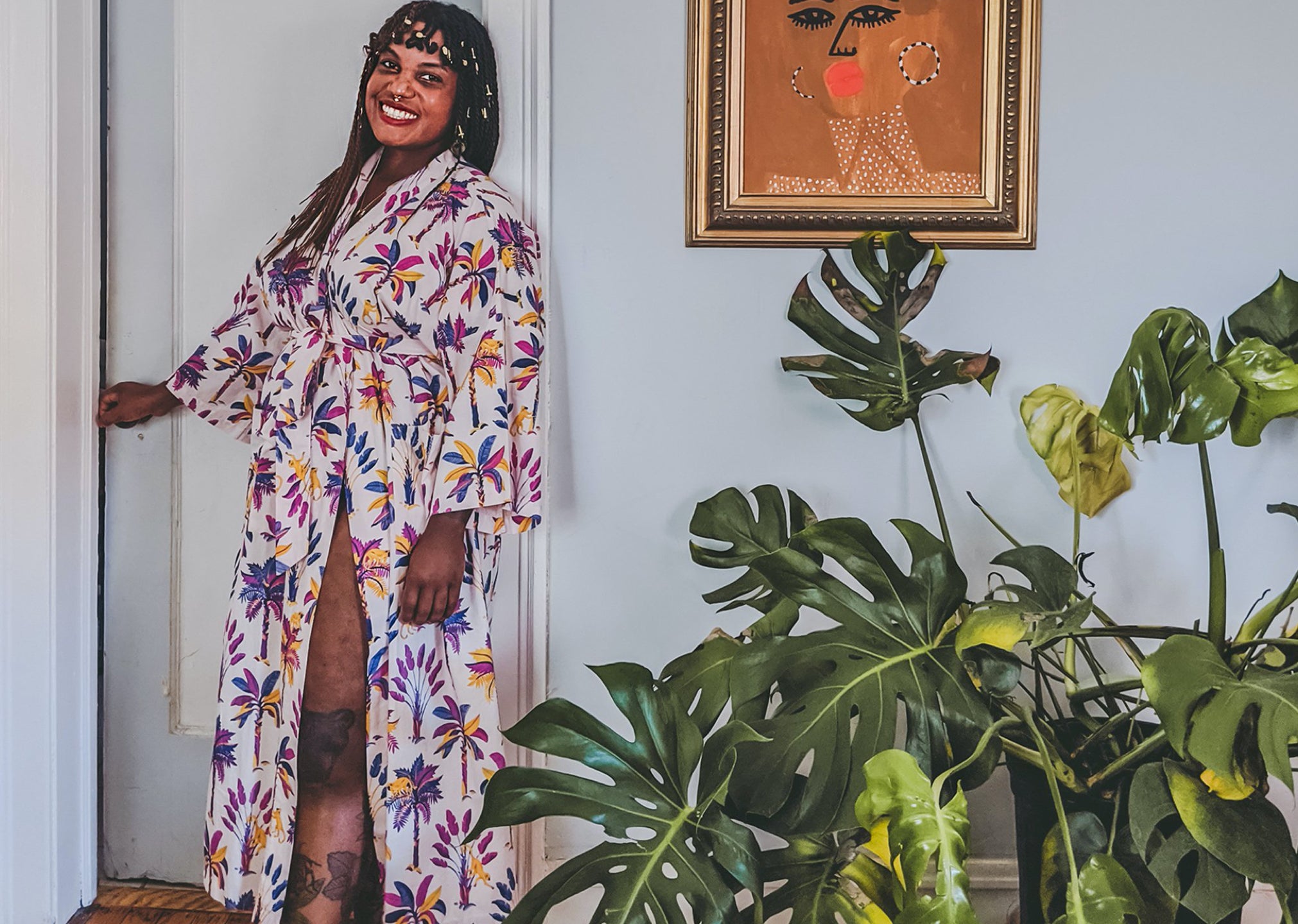 Smiling person in a floral robe standing beside a white door and leafy houseplants